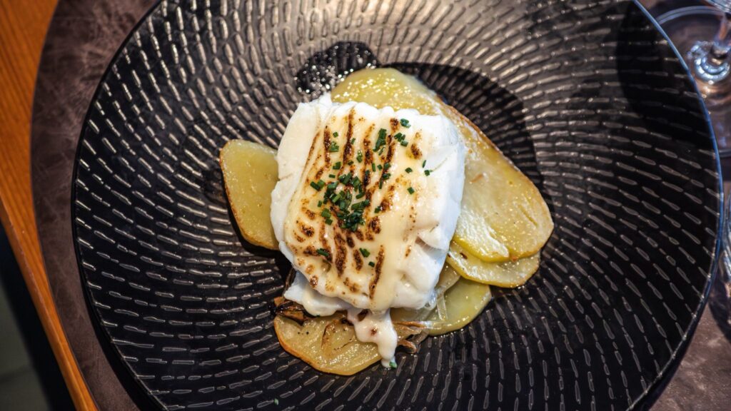 Plato negro con hendiduras con patatas cortadas en láminas y bacalao gratinado con alioli de Ses Oliveres