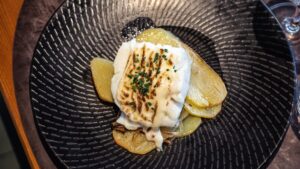 Plato negro con hendiduras con patatas cortadas en láminas y bacalao gratinado con alioli de Ses Oliveres